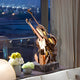 The modern artistic decor showcases the Harmonic Copper Violin Steel Sculpture (80cm) by Giant Sculptures. This stunning sculpture, with a metallic finish on a marble base, is placed indoors near a window where night city lights shimmer. Beside it sits a table adorned with white and yellow flowers.