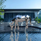 A large Horse Geometric Steel Outdoor Sculpture from Giant Sculptures stands over a water feature with a waterfall backdrop. Its mirrored surface reflects the surrounding water and sky, while lush greenery enhances the tranquil setting.