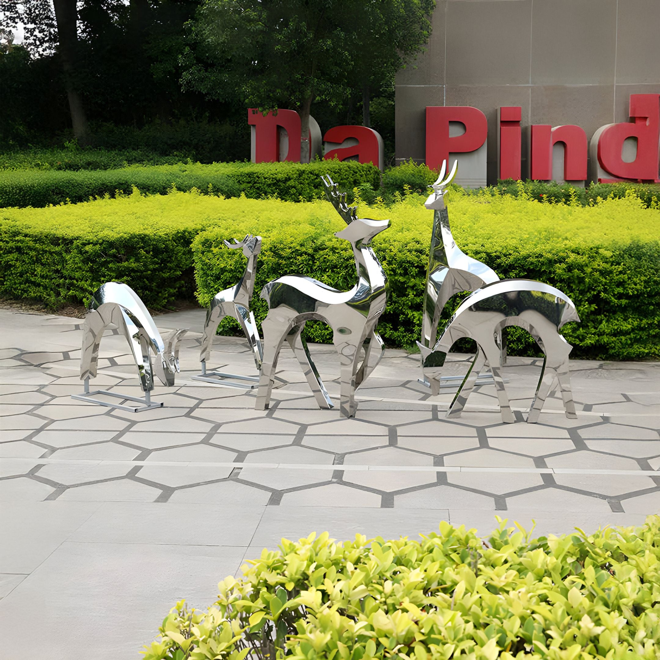 Four Deer Organic Steel Outdoor Sculptures by Giant Sculptures, exuding modern artistry, grace a patterned pavement before trimmed hedges and partial red signage. The backdrop of trees enhances the allure of this stainless steel installation.