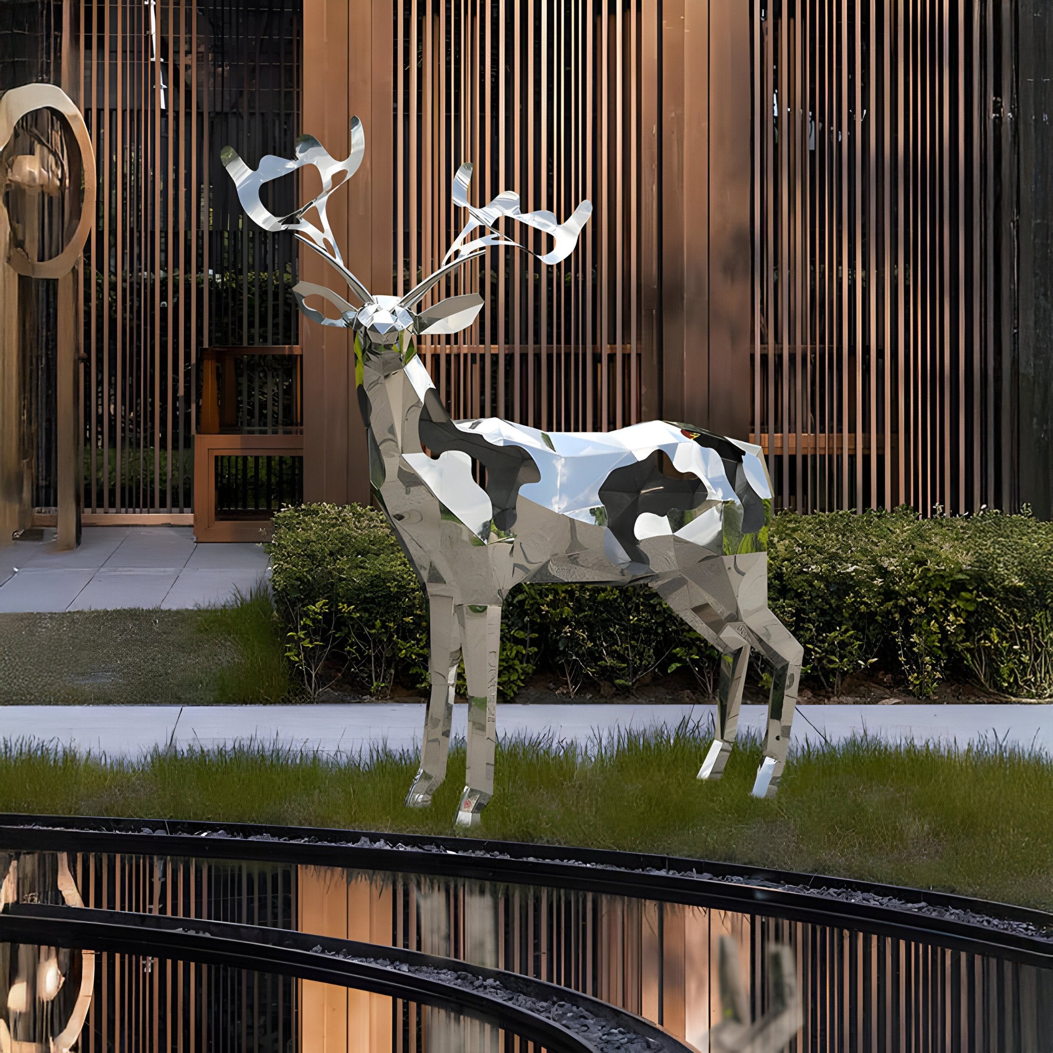 In a manicured garden, the Mystic Deer Geometric Steel Outdoor Sculpture from Giant Sculptures stands elegantly. This 197cm stainless steel piece features a reflective surface with abstract patches that beautifully catch the light. A neatly trimmed hedge and vertical wooden slats create a striking backdrop, while a circular fountain adds to the sophistication of this outdoor décor.