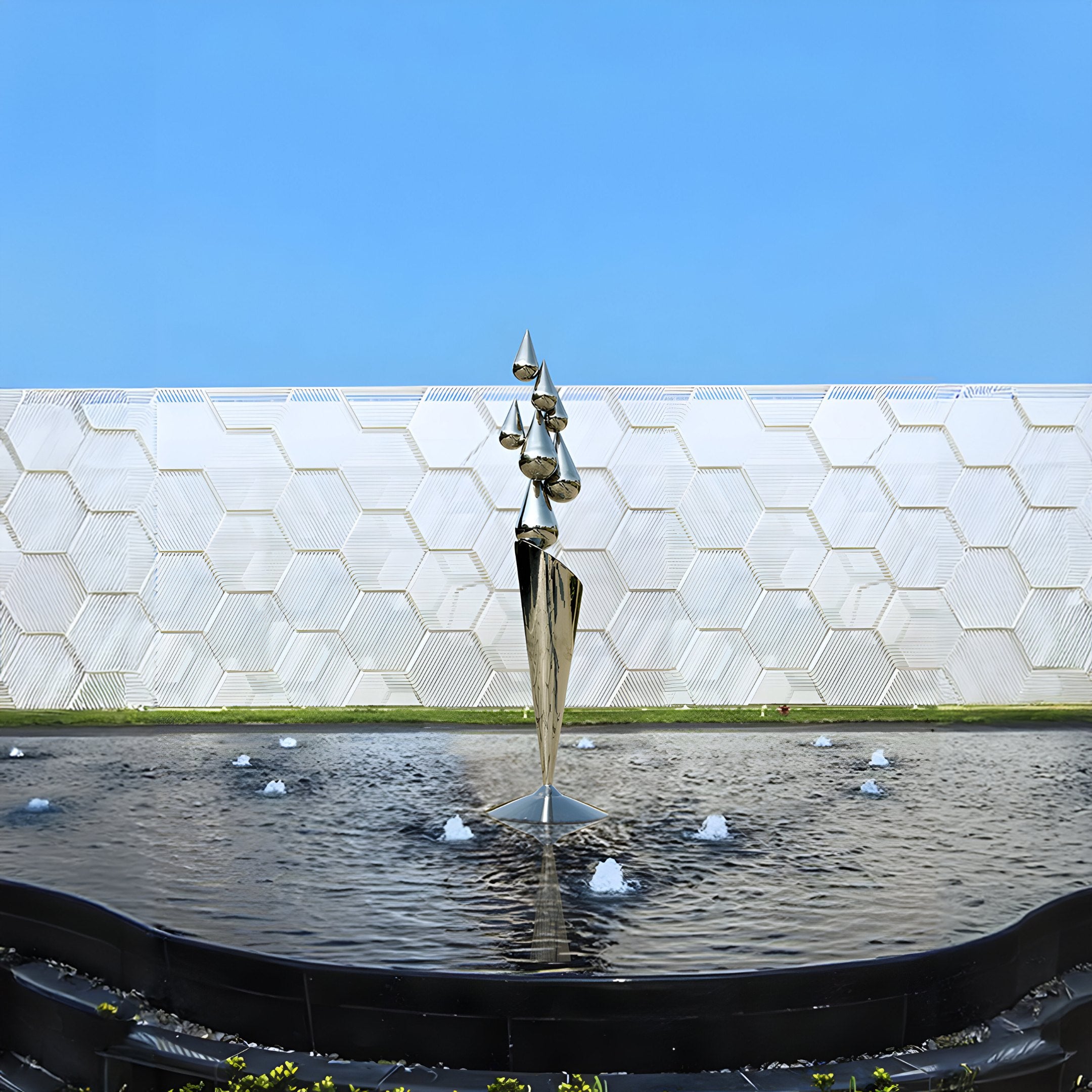 The Teardrop Abstract Steel Outdoor Sculpture by Giant Sculptures stands 215 cm tall in a circular fountain with small jets, set against a large wall featuring a geometric honeycomb pattern under a clear blue sky.