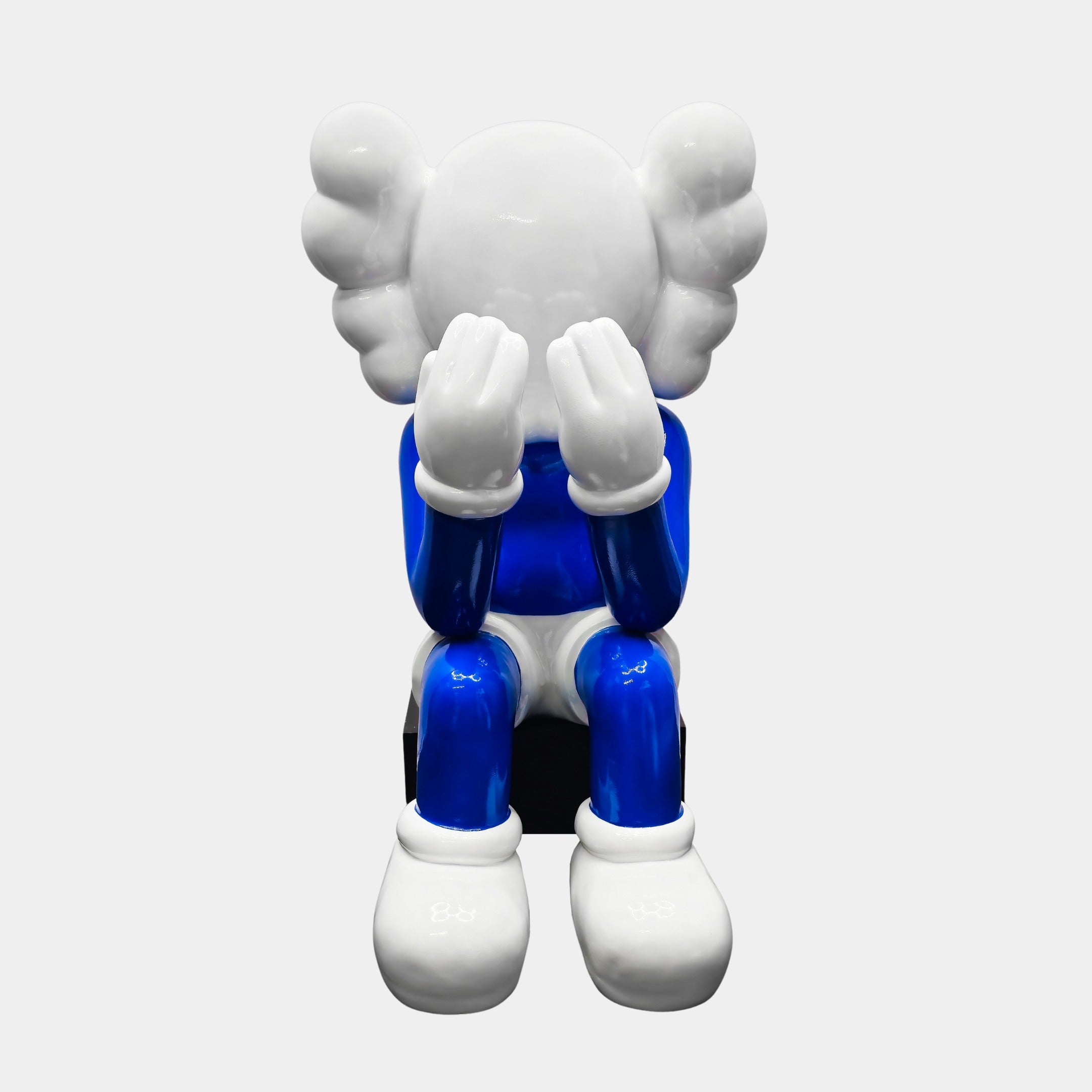 The Ex-Display White & Blue Iconify Melancholy Sitting Sculpture by Giant Sculptures, features a stylized figure with a round face and cloud-like ears. It sits with hands on its face, cartoon-like shoes, painted in blue and white—a perfect centerpiece for minimalist design enthusiasts.