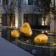 A nighttime scene showcasing modern art installations: three "Ripple Gold Organic Outdoor Steel Sculptures" by Giant Sculptures, featuring a sleek gold finish with black stripes, are positioned above a reflective water surface. The sculptures are surrounded by illuminated trees and a contemporary building in the background.