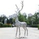 A Giant Sculptures Luminous Stag Silver Steel LED Sculpture, standing 212cm tall, with intricate metal patterns and slender antlers, graces the pavement. It is crafted from silver stainless steel and set against trees and city buildings under a cloudy sky.