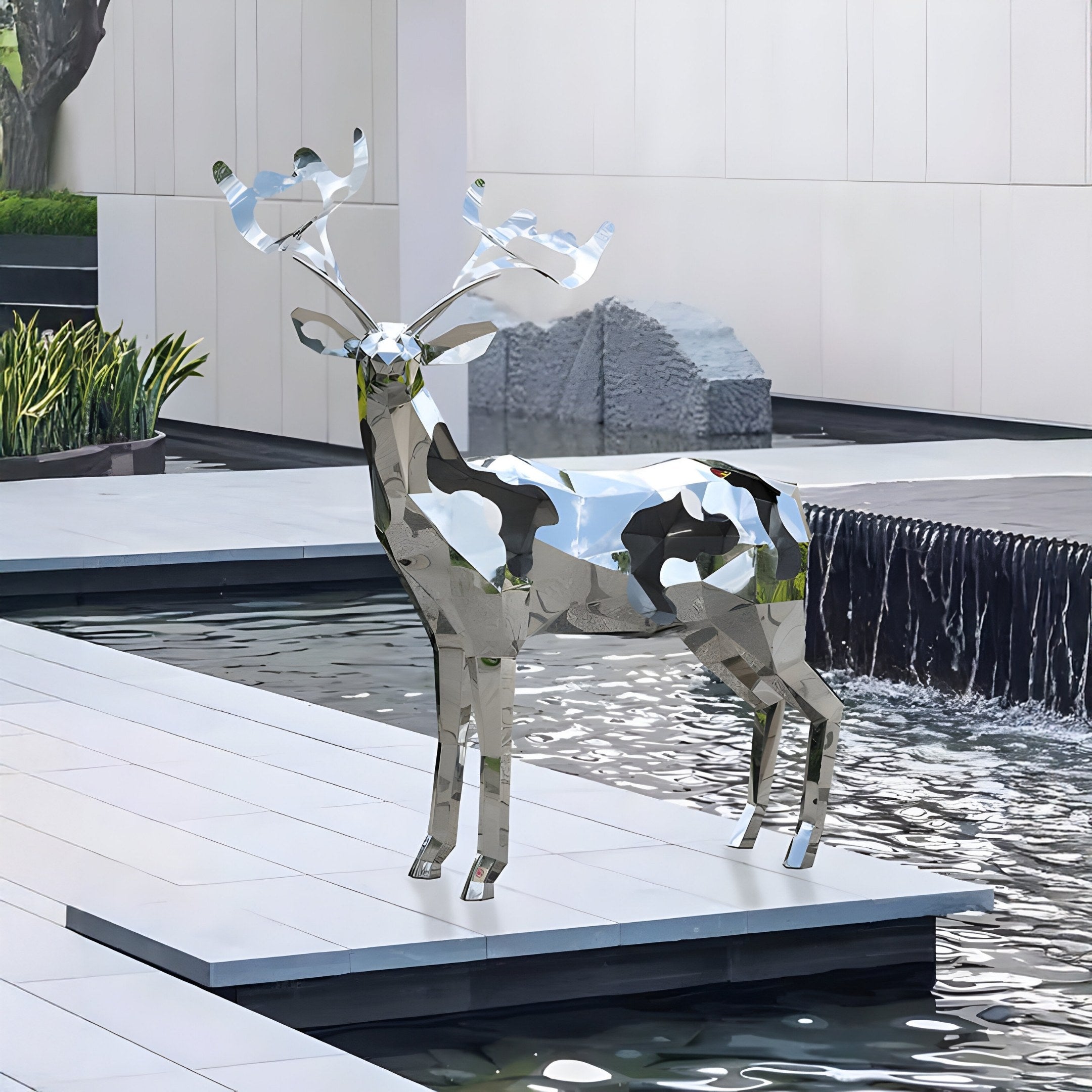 The Mystic Deer II Geometric Steel Outdoor Sculpture from Giant Sculptures, standing at 197cm, is showcased on a platform by a modern water feature with cascades. A minimalist garden with plants and a large stone against a white building enhances its allure.