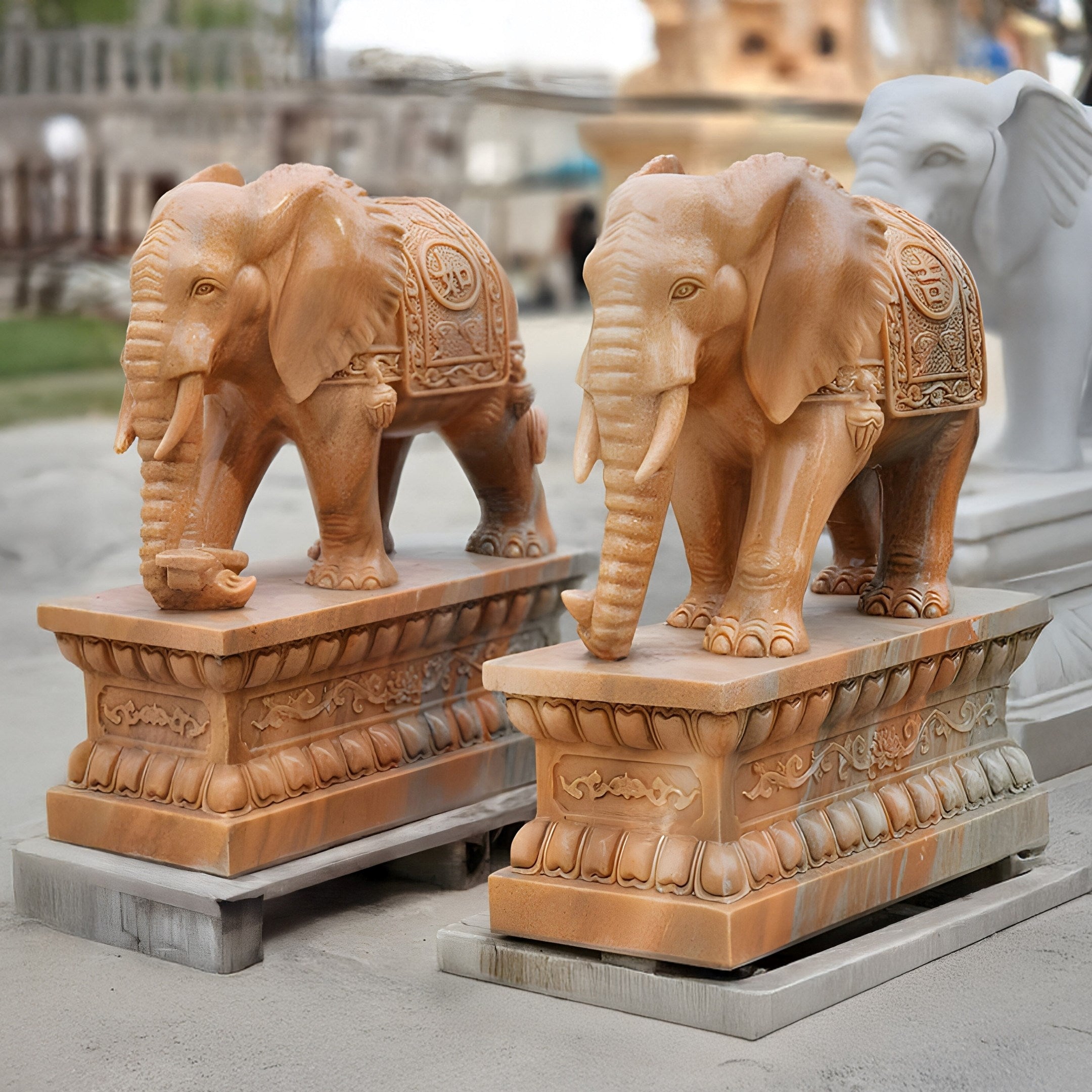 Two giant yellow marble elephant sculptures, intricately hand-carved and adorned with ornate designs stand outdoors under a partly cloudy sky. The primary focus is on the exquisite Elephant Yellow Marble Outdoor Sculpture - 180cm by Giant Sculptures, with a third blurred statue in the background.