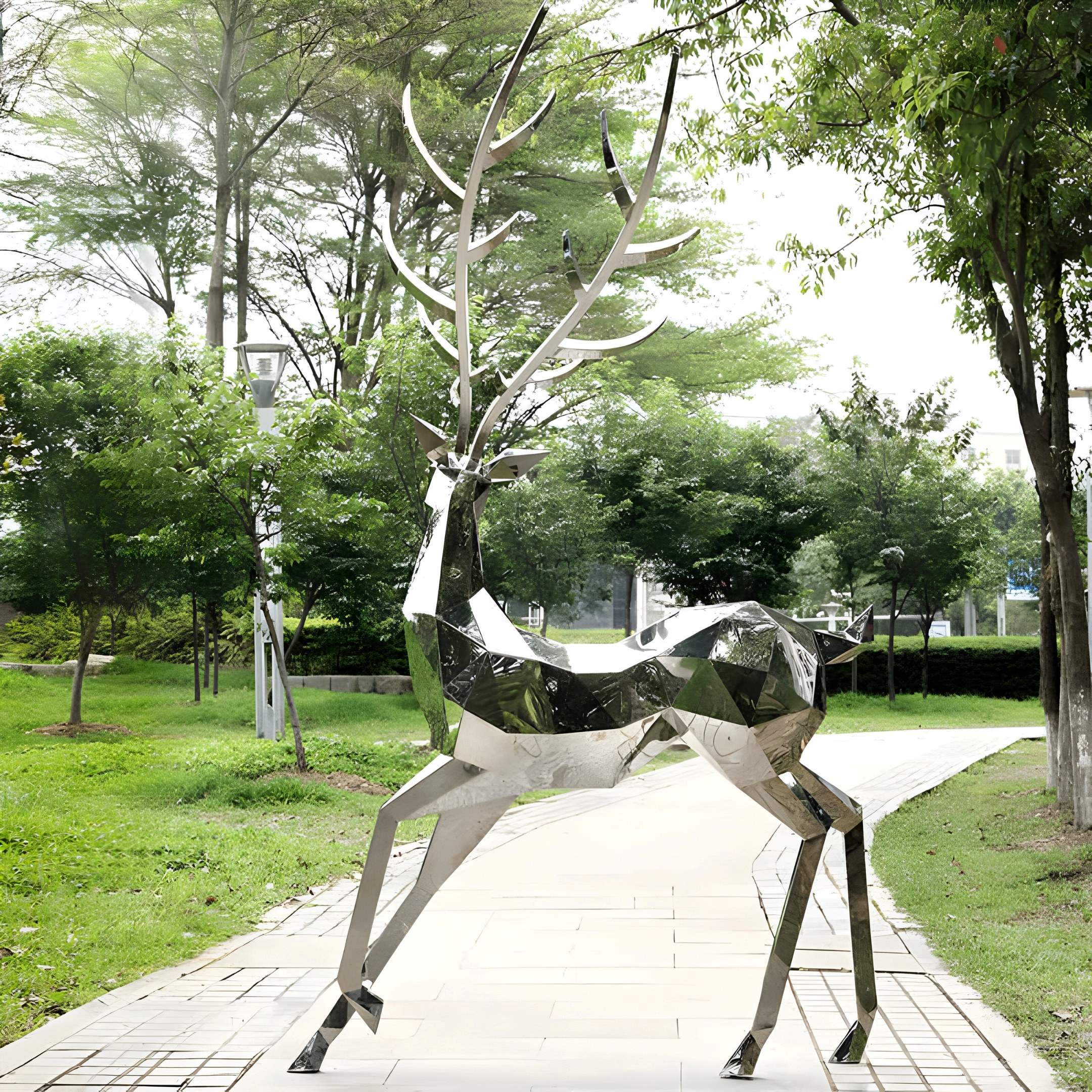 The Noble Deer IV Geometric Steel Sculpture by Giant Sculptures towers on a paved path, embraced by verdant trees and grass. The 258cm stainless steel artwork reflects light beautifully amid leafy foliage and a clear sky.