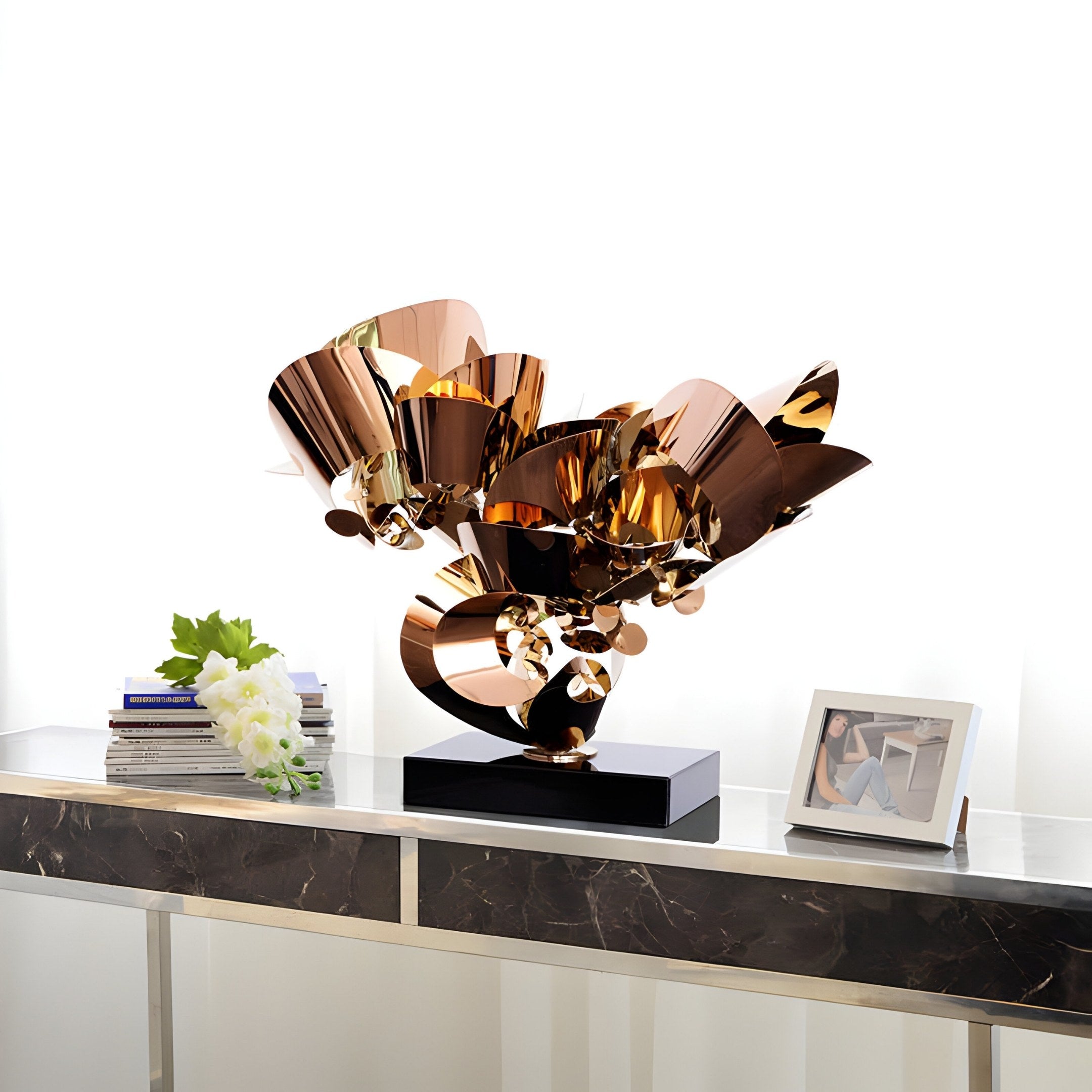 The Petal Fusion Copper Organic Steel Sculpture - 70cm by Giant Sculptures features modern, shiny, curved surfaces on a black base. Displayed on a marble console table with books, white flowers, and a framed photo, the soft white background enhances its elegant composition.