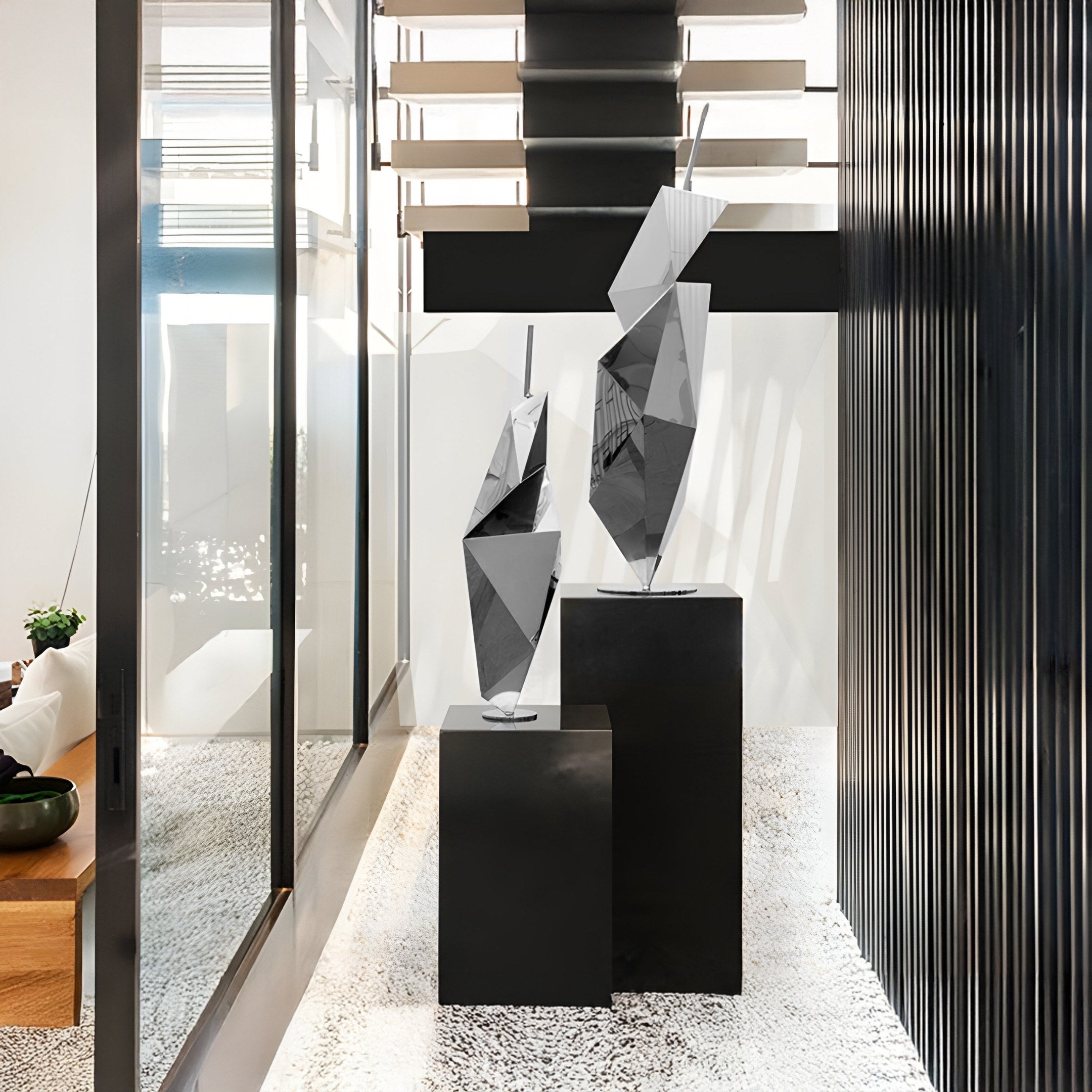 In a modern, well-lit corridor with a speckled floor and slatted wooden above, two Prism Silver Geometric Steel Sculptures by Giant Sculptures stand on black pedestals. A large window on the left enhances the contemporary art ambiance.