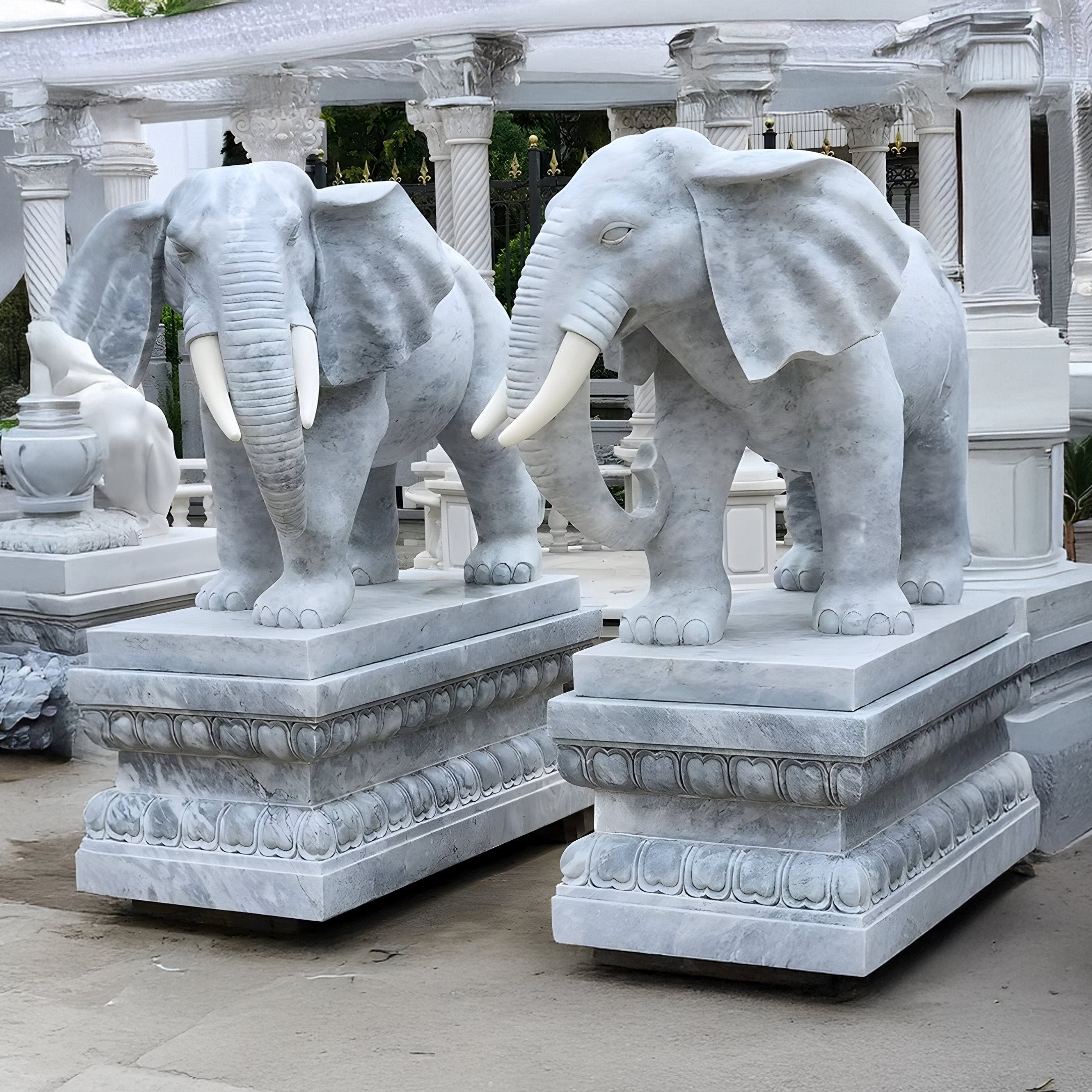 Two large Elephant Grey Marble Outdoor Sculptures by Giant Sculptures, each 120cm and with prominent tusks, stand on intricately carved pedestals. The backdrop includes elegant white columns and architectural details, suggesting an outdoor display or garden setting.