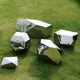 Five Tetra Geometric Steel Rock Sculptures by Giant Sculptures are scattered on a grassy field. These contemporary pieces have angular shapes and mirrored surfaces that reflect their surroundings, including trees and buildings.