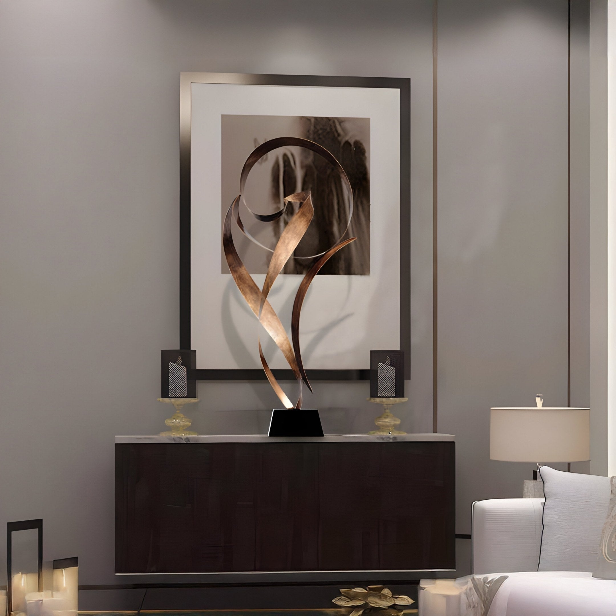 A modern living room features the Ribbon Bronze Abstract Steel Sculpture by Giant Sculptures on a dark wood console table beneath framed art. Two decorative candle holders flank it, and a white sofa with a tall lamp is partially visible to the right.