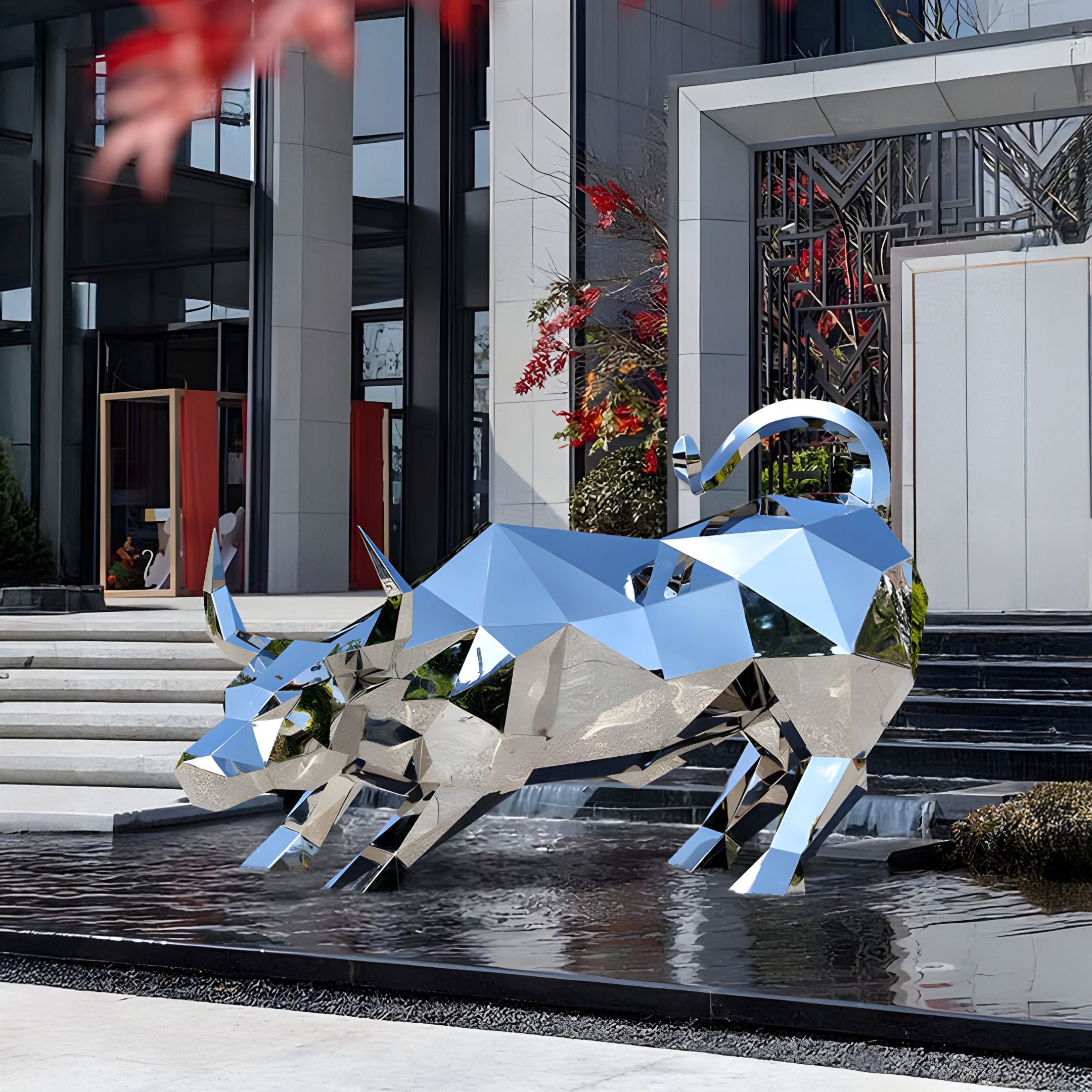 A stunning geometric steel sculpture named "Toro Bull," crafted by Giant Sculptures, is prominently displayed atop a water feature in front of a contemporary building with expansive windows and stairs. The vibrant red foliage and plants serve as a beautiful backdrop to this striking outdoor piece, which stands at 193cm tall.