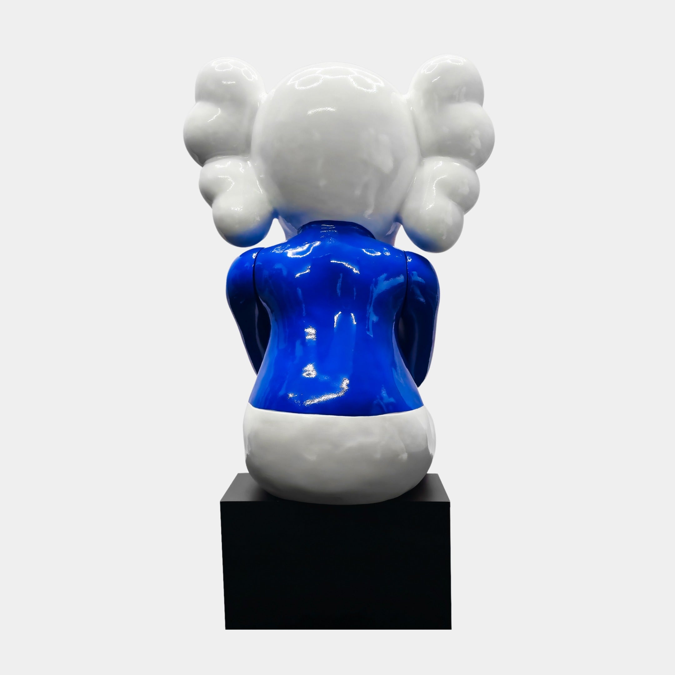 The Ex-Display White & Blue Iconify Melancholy Sitting Sculpture by Giant Sculptures is a contemporary piece featuring a cartoonish figure on a black pedestal with a minimalist design. It has a glossy finish, white head with cloud-like ears, blue body, and white lower half—ideal as a modern centerpiece.