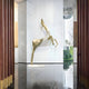 The Deer Gold Geometric Wireframe Outdoor Sculpture by Giant Sculptures elegantly stands in a minimalistic room with marble walls and glossy black floors. Vertical wooden panels complement its design while a large window showcases the lush greenery outside.