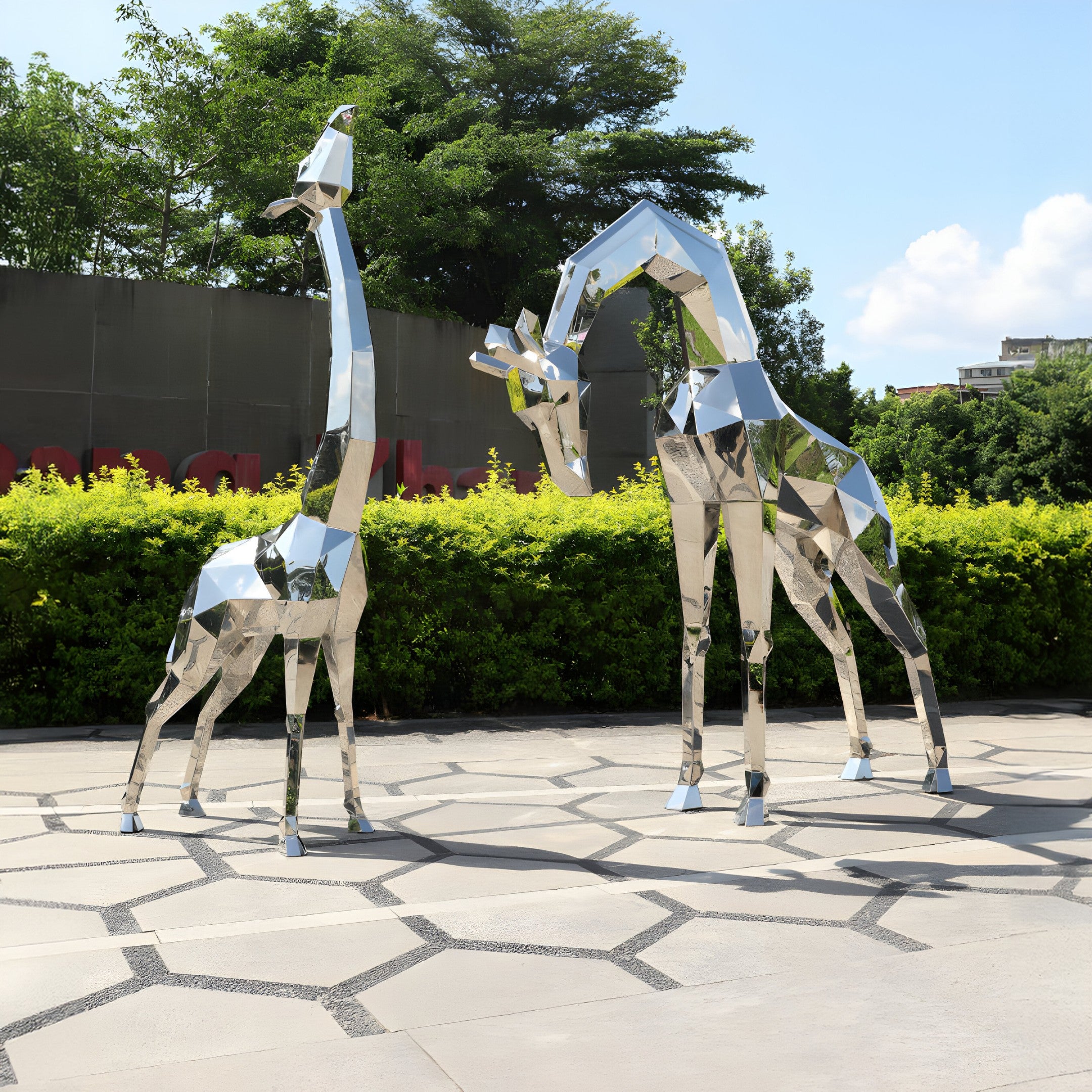 The Giraffe II Geometric Steel Outdoor Sculpture by Giant Sculptures stands gracefully at 200cm, showcasing a striking example of geometric design with its angular, faceted surface. Positioned on a paved area, the larger sculpture bends its neck towards a smaller one, while green shrubs and trees create a serene backdrop under a clear blue sky. This scene elegantly captures the essence of contemporary art.