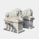 The Giant Lying Lion Marble Outdoor Sculptures from Giant Sculptures rest elegantly on stone pedestals, epitomizing sophisticated garden decor. The lions' intricately detailed manes and commanding expressions are captivating, while the vertical grooves on the pedestals create intriguing shadows against a plain, bright backdrop.