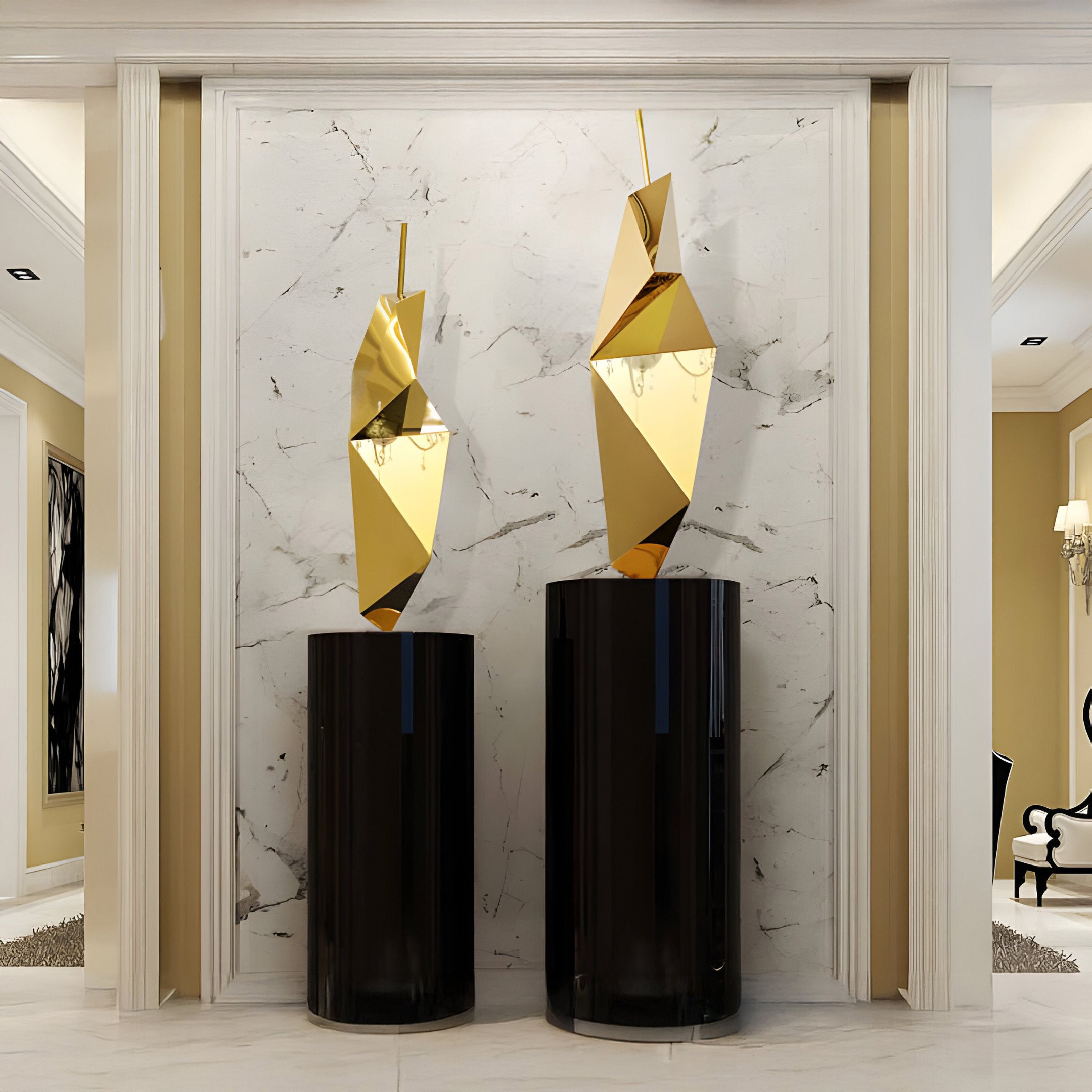 Two Prism Gold Geometric Steel Sculptures by Giant Sculptures, each 80cm tall and resembling penguins, are perched on tall black cylindrical pedestals against a white marble wall with gray veins. The arrangement exudes contemporary design elegance, enhanced by warm lighting in a modern interior.