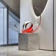 The Ribbon Red III Abstract Steel Sculpture by Giant Sculptures, featuring intertwined red and silver metallic ribbons, stands elegantly on a stone pedestal within a modern interior with glass elements and a mirrored wall.