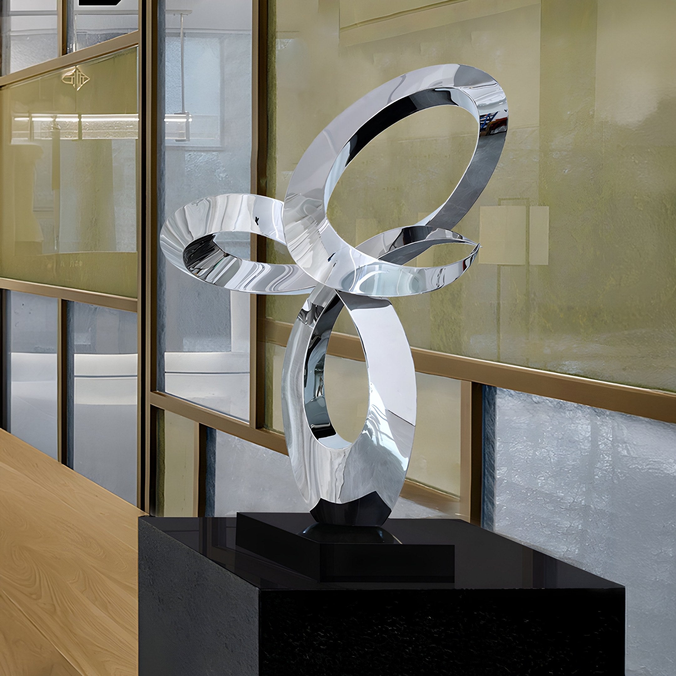 The Infinity Loop Abstract Steel Sculpture by Giant Sculptures stands elegantly at 105cm, featuring interlocking polished metallic loops on a black pedestal. Bathed in natural light through large windows, it contrasts with glass and beige panels for a modern artistic display.