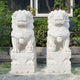 In front of a building with lush greenery, two Chinese Guardian Lion IV Marble Outdoor Sculptures from Giant Sculptures stand side by side. Each 180cm statue showcases intricate carvings on its pedestal, set against a window and various plants to enhance the scenes elegance.