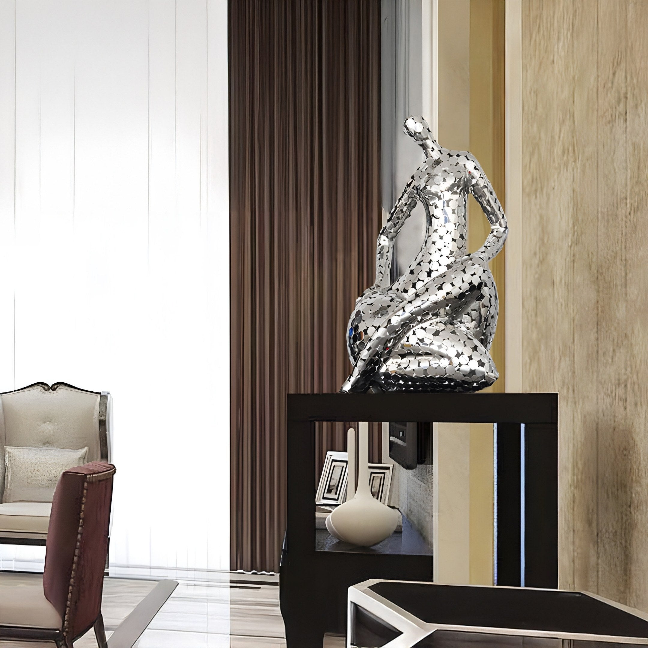 A modern interior features the Solace Silver Abstract Steel Sculpture by Giant Sculptures on a black pedestal. Alongside a cushioned armchair and sleek coffee table against a textured beige wall, the space exudes tranquility and contemporary elegance.