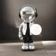 The Lunar Silver Astronaut Glow Orb Sculpture (80cm) by Giant Sculptures features a shiny astronaut in a silver jacket with a metallic helmet, holding a glowing orb. It stands on wood with ambient lighting enhancing its modern, futuristic feel.