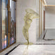 A sleek hallway with marble floors and walls features the Tornado Gold Organic Steel Sculpture - 152cm by Giant Sculptures. Resembling a twisting, elongated figure, it captivates as sunlight filters through large metal-framed windows lining the left side, enhancing this extraordinary artwork.