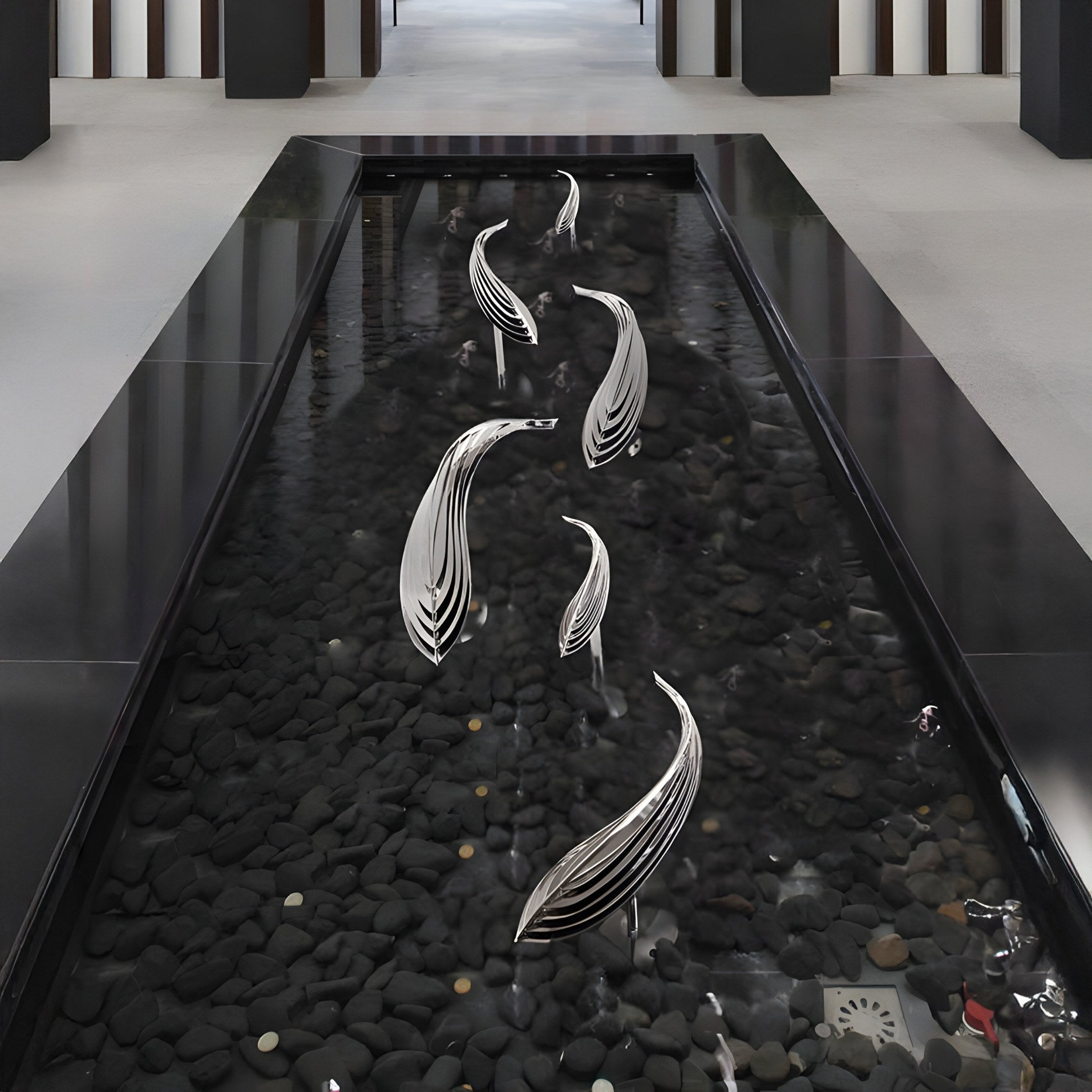 The indoor water feature highlights Giant Sculptures Organic Fish Abstract Steel Sculptures over dark stones. The sculptures appear to float above the shallow water amidst a minimalist black and white decor.