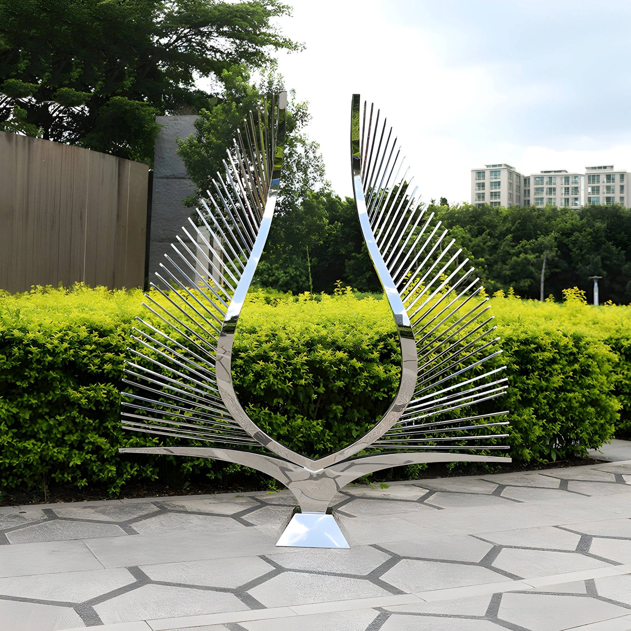 A 200cm Celestial Rays Steel Outdoor Sculpture by Giant Sculptures stands on a tiled path, with green bushes and trees behind it. The stainless steels sleek design contrasts with nature, as tall buildings rise in the contemporary landscapes background.