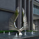 A sleek, modern sculpture crafted from steel and designed to resemble abstract wings stands majestically on a pedestal within a shallow fountain. The Celestial Rays Steel Outdoor Sculpture by Giant Sculptures, measuring 200 cm in height, is prominently displayed in front of a contemporary building adorned with large windows and vertical panels, with small fountains elegantly surrounding its base.