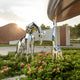 In a lush garden adorned with pink flowers, the Giraffe II Geometric Steel Outdoor Sculpture by Giant Sculptures stands 200cm tall alongside its calf. In the background, a curved modern building with a geometric facade features a sign that reads ras, all beneath a cloudy sky.