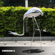 A Giant Sculptures Crane Contemporary Outdoor Steel Sculpture - 98/158cm stands on a sidewalk with greenery, featuring an arched neck and legs for a modern art elegance. A VERSION 4 label is in the bottom left corner.