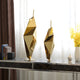 A pair of Prism Gold Geometric Steel Sculptures from Giant Sculptures grace a dark marble console table in front of a bright window. Yellow flowers in a vase and a stack of books enhance the contemporary design, resulting in an elegant display.