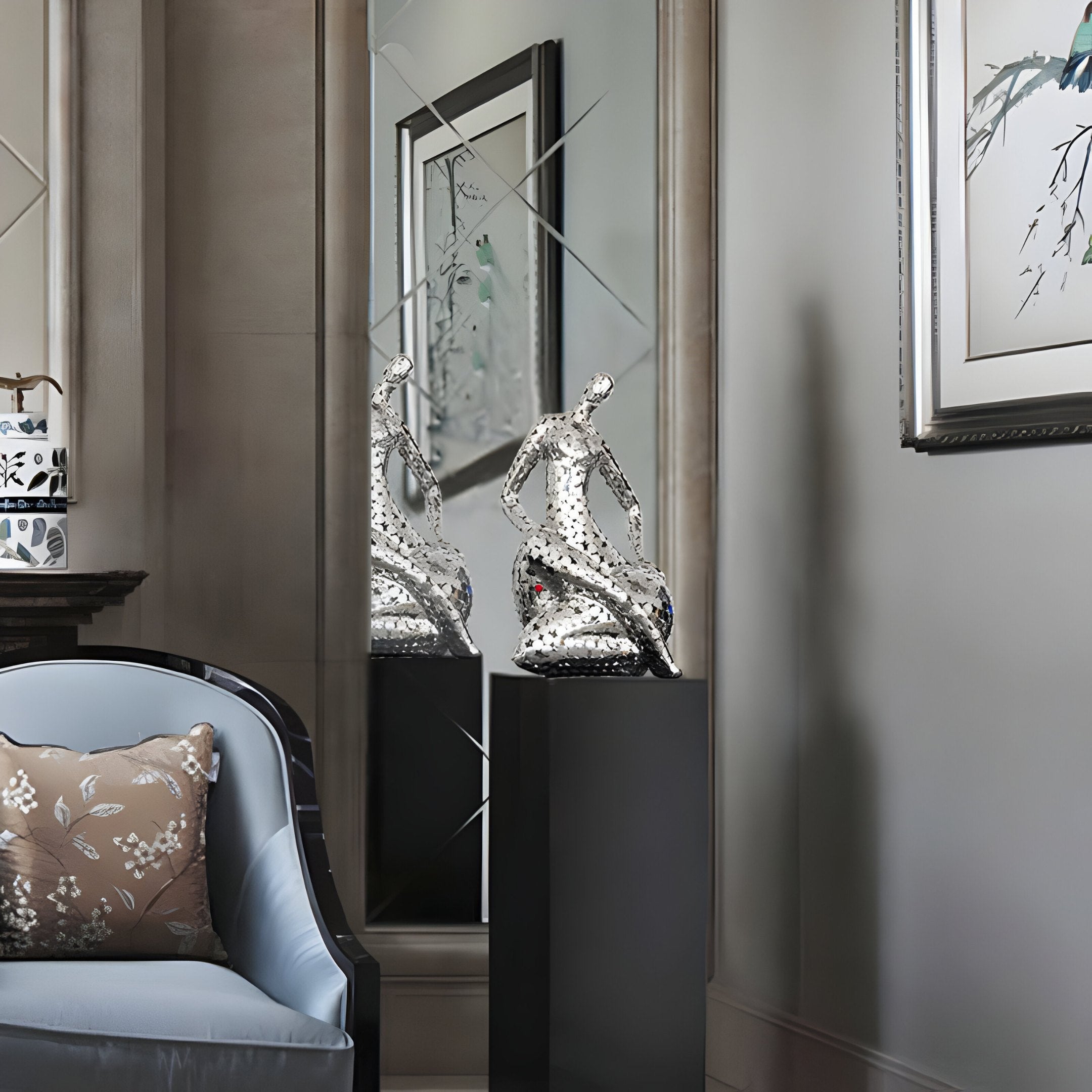 The Solace Silver Abstract Steel Sculpture - 81cm by Giant Sculptures elegantly stands on a black pedestal against a mirrored wall, featuring an abstract human figure in a seated position. The sophisticated room is adorned with modern abstract art, framed pieces, a plush chair accented with a floral pillow, and subtle lighting.