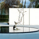 The "Magical Antler Steel Outdoor Sculpture - 235cm" by Giant Sculptures depicts a modern stainless steel deer with enchanting antlers rising from a reflective circular water pool. The antlers feature flat, disc-like shapes, and the scene is completed by a white wall, tall trees, and a partially visible building in the background.