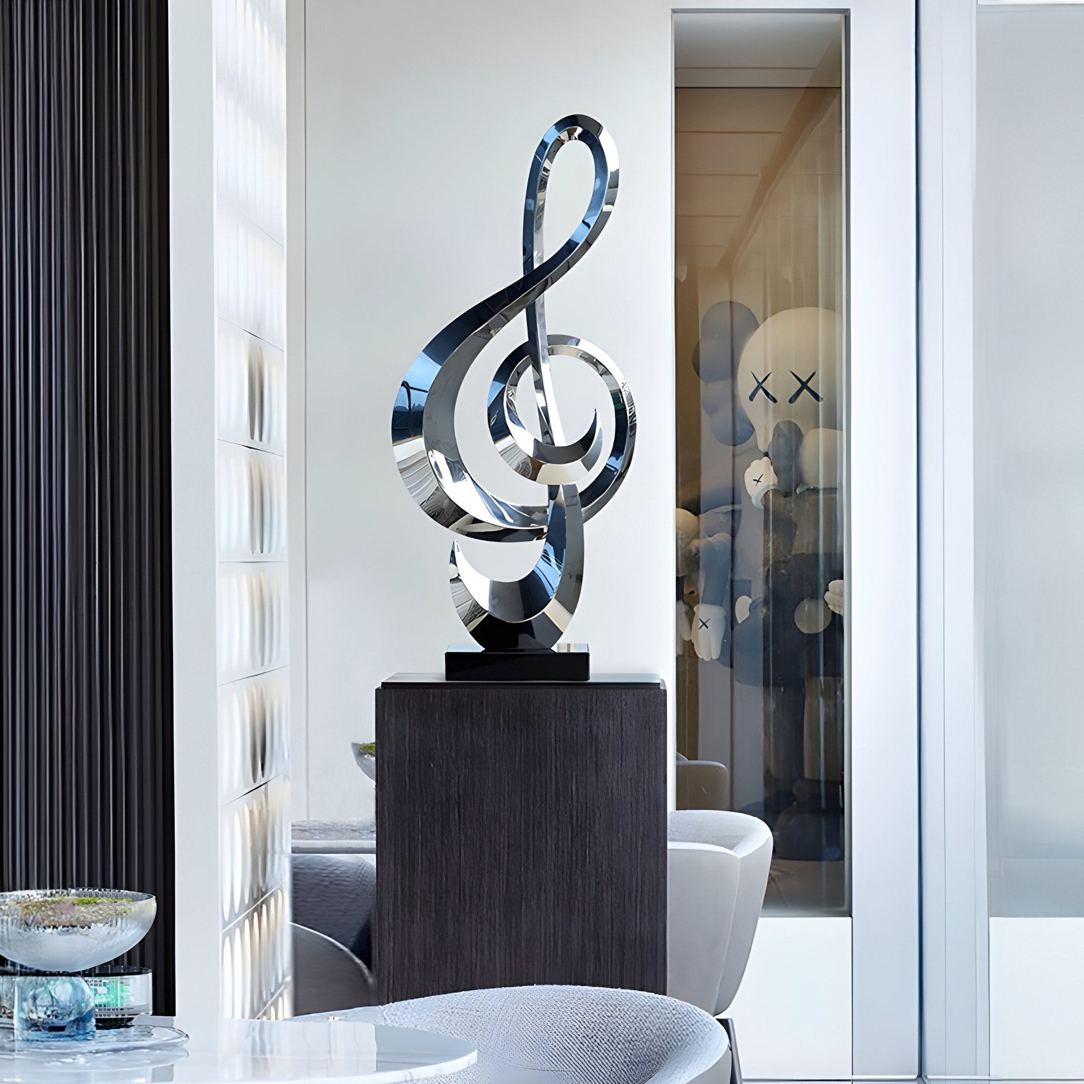 A modern interior showcases the Melodic Abstract Steel Sculpture - 106cm by Giant Sculptures, elegantly perched on a dark pedestal. In the background, a contemporary room with light walls and abstract art featuring X-eyed characters is visible through the glass partition.