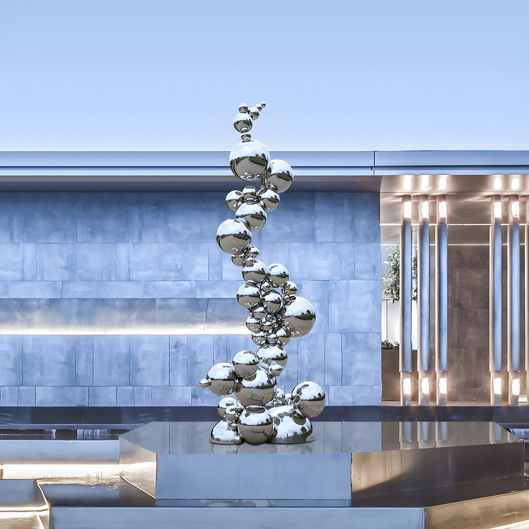 The Molecular Silver Organic Steel Outdoor Sculpture by Giant Sculptures is crafted from reflective spheres in stainless steel, stacked on a metallic pedestal. It stands gracefully in a contemporary setting with gray walls and vertical light panels. Height: 226cm.