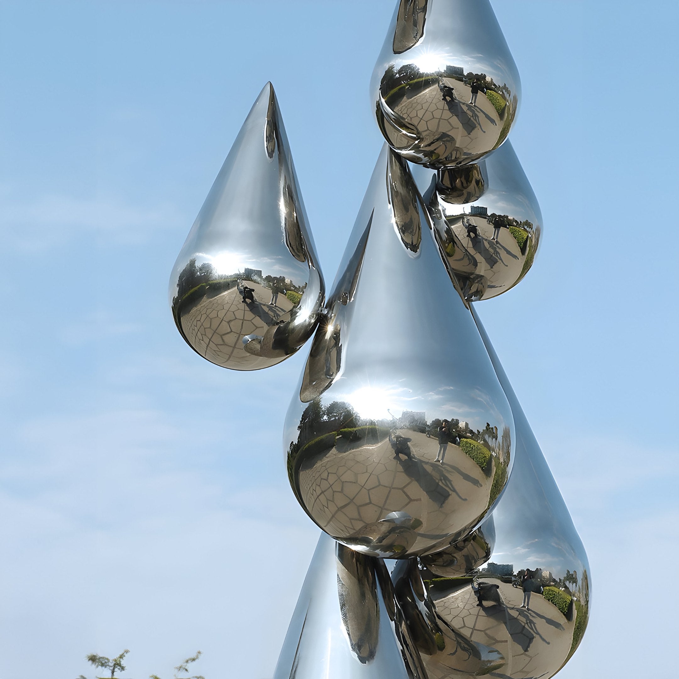 The Teardrop Abstract Steel Outdoor Sculpture by Giant Sculptures stands tall at 215cm, gleaming against the blue sky. Its reflective surface creates mirror-like images of its surroundings and people, transforming them into captivating abstract forms.