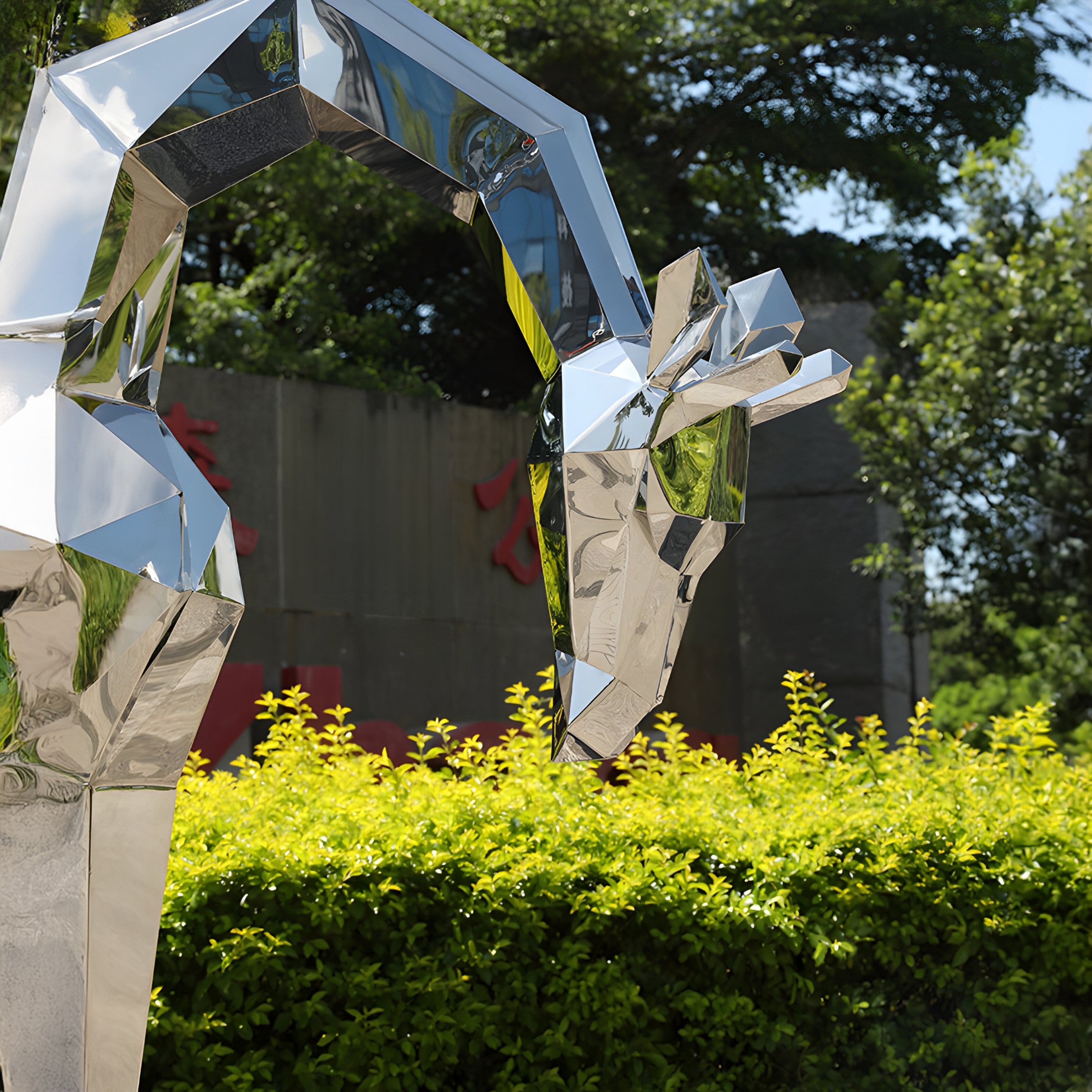 A Giraffe Geometric Steel Outdoor Sculpture, standing 181 cm tall and crafted by Giant Sculptures, gleams amidst vibrant green bushes. This modern outdoor piece boasts an angular design. In the background, trees and a large dark wall with red lettering provide contrast, while sunlight brightly illuminates the scene.