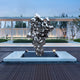 The Molten Silver Organic Steel Sculpture (155cm) by Giant Sculptures, an abstract stainless steel piece with a reflective surface, stands on a white platform outdoors. The backdrop features trees and a glass-walled building under clear skies, with pathways and outdoor lights enhancing the scene.