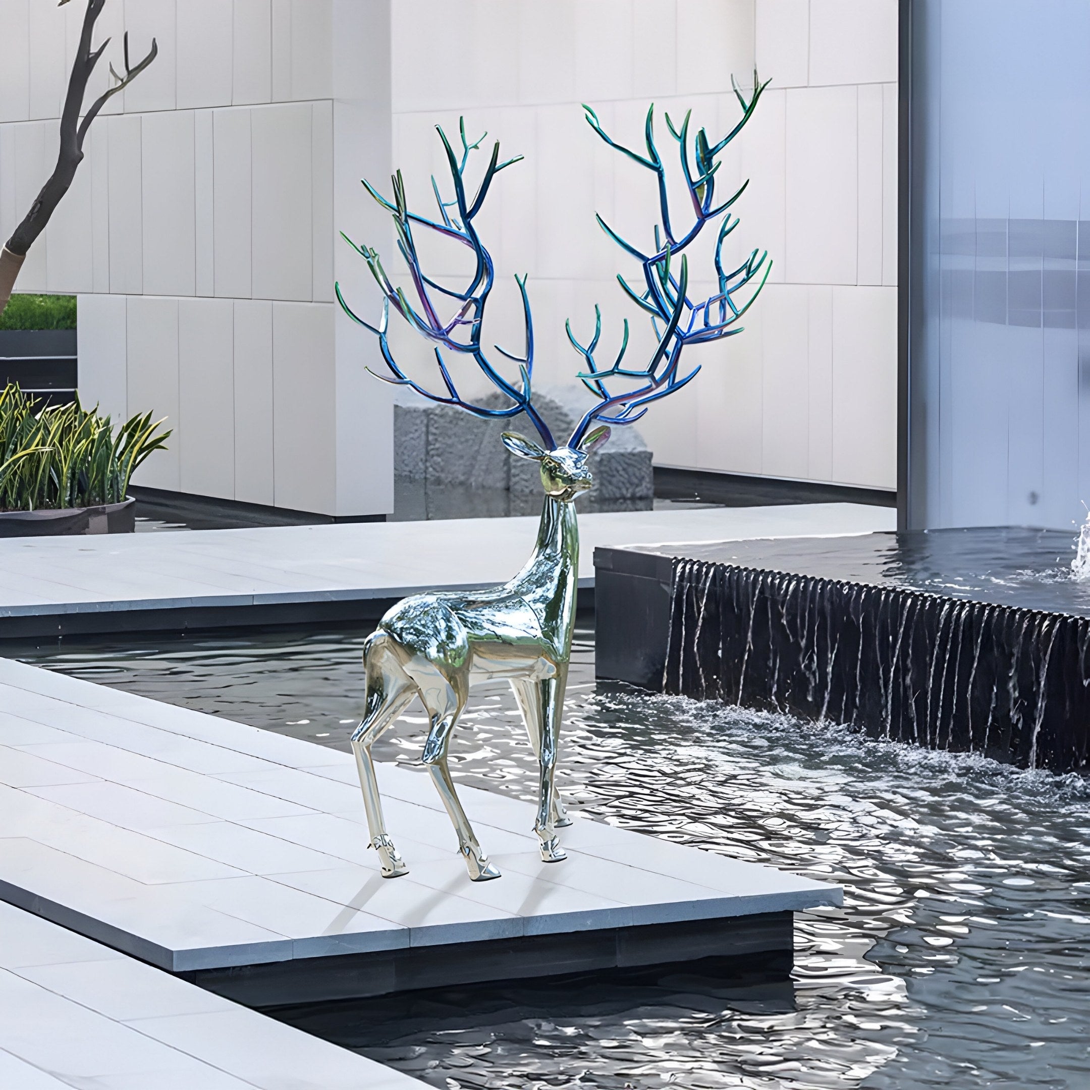 The Mystic Stag Steel Outdoor Sculpture by Giant Sculptures features shiny stainless steel and multicolored antlers, standing on a white platform beside a reflecting pool and small waterfall in a modern outdoor setting, showcasing art amid geometric architectural elements.