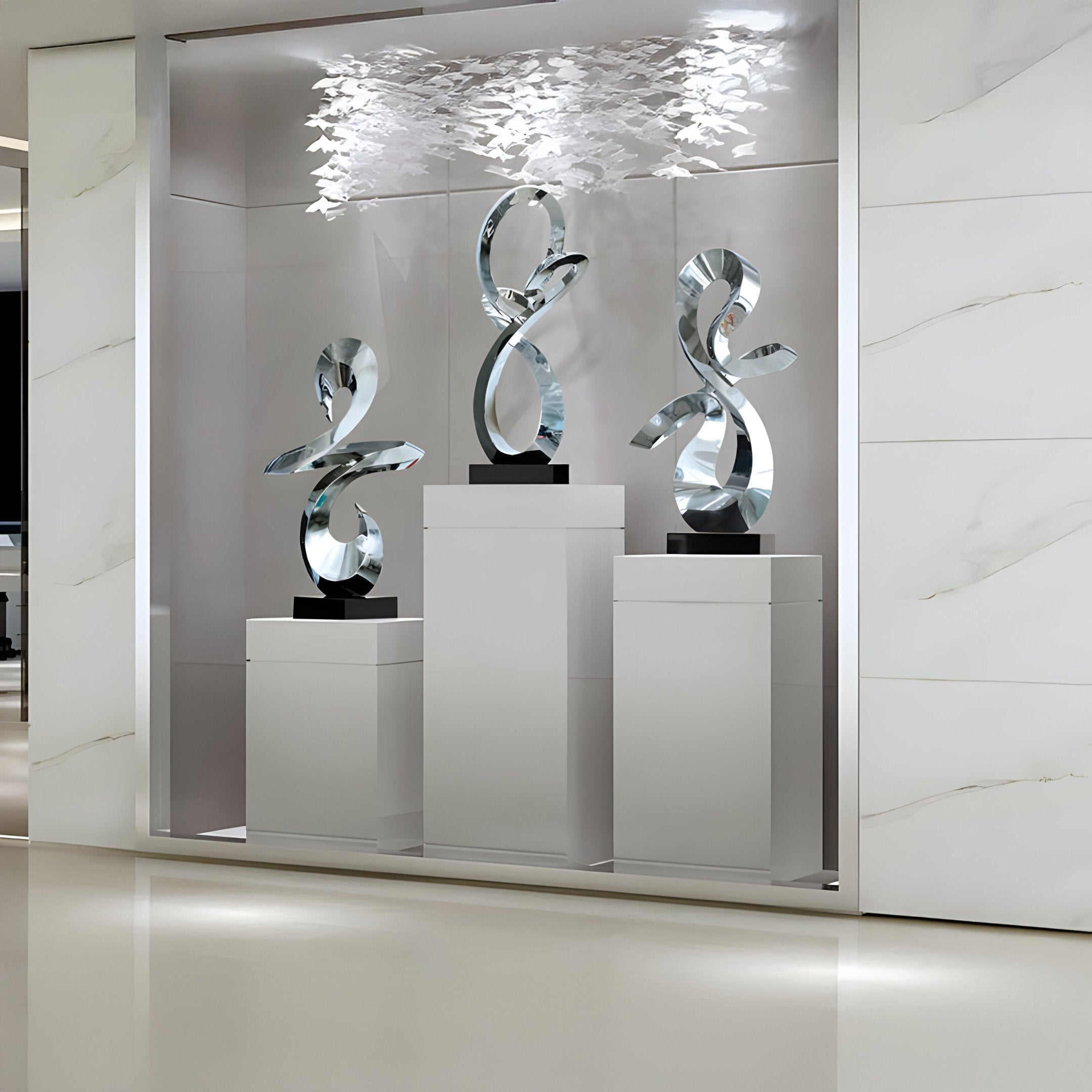 A display showcasing three "Serpentine Silver Abstract Steel Sculptures" by Giant Sculptures stands on white pedestals in a modern gallery setting. Above them, feather-like decorations dangle from the ceiling, gently highlighted by soft lighting. The walls and floor maintain a minimalistic and light-colored ambiance.