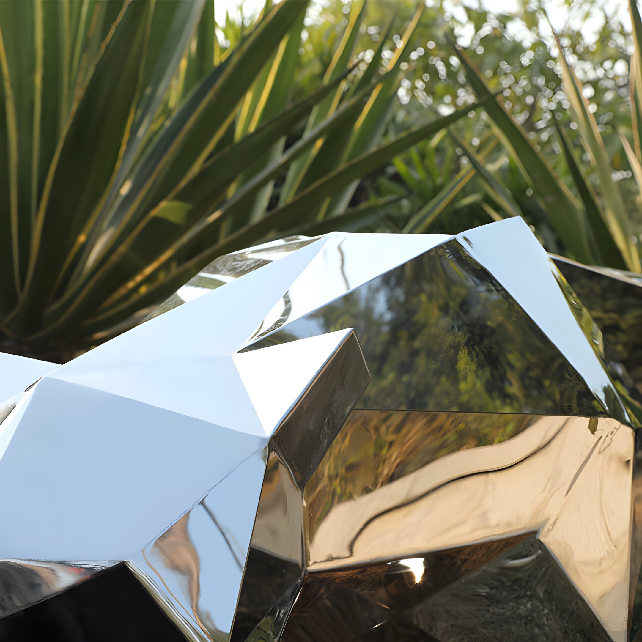 A close-up of the Panther II Geometric Steel Outdoor Sculpture by Giant Sculptures showcases its reflective metallic surface nestled among verdant, spiky-leaved plants. This outdoor piece mirrors the vibrant greenery and light, creating a complex interplay of reflections reminiscent of a panther's stealth.