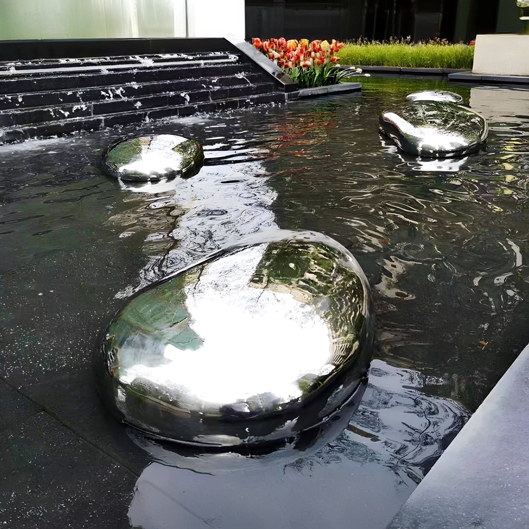 Giant Sculptures Pebble Organic Outdoor Steel Sculptures (90/118cm) create a modern landscape as they sit in a shallow pool, reflecting the sky and nearby buildings. Red tulips in the background add a colorful touch to this artistic display.