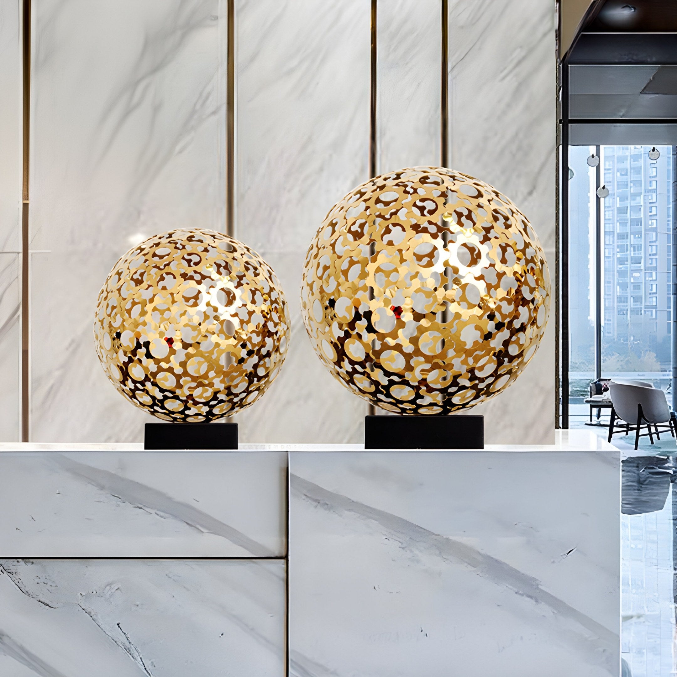 Two Eclipse Gold Abstract Steel Sculptures by Giant Sculptures, showcasing intricate designs, elegantly adorn a marble countertop. Large marble panels and tall windows behind them offer a view that resembles the graceful fusion of modern sculpture and urban elegance.