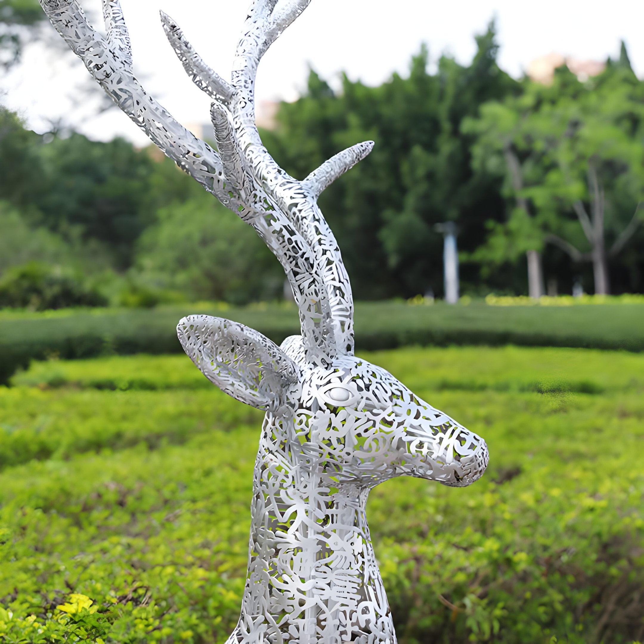 A detailed view of the Luminous Stag Silver Steel LED Sculpture by Giant Sculptures reveals its elegant silver stainless steel craftsmanship, intricate cut-out patterns, and majestic antlers. This eye-catching piece stands gracefully at 212 cm against a lush garden backdrop filled with trees and shrubs.