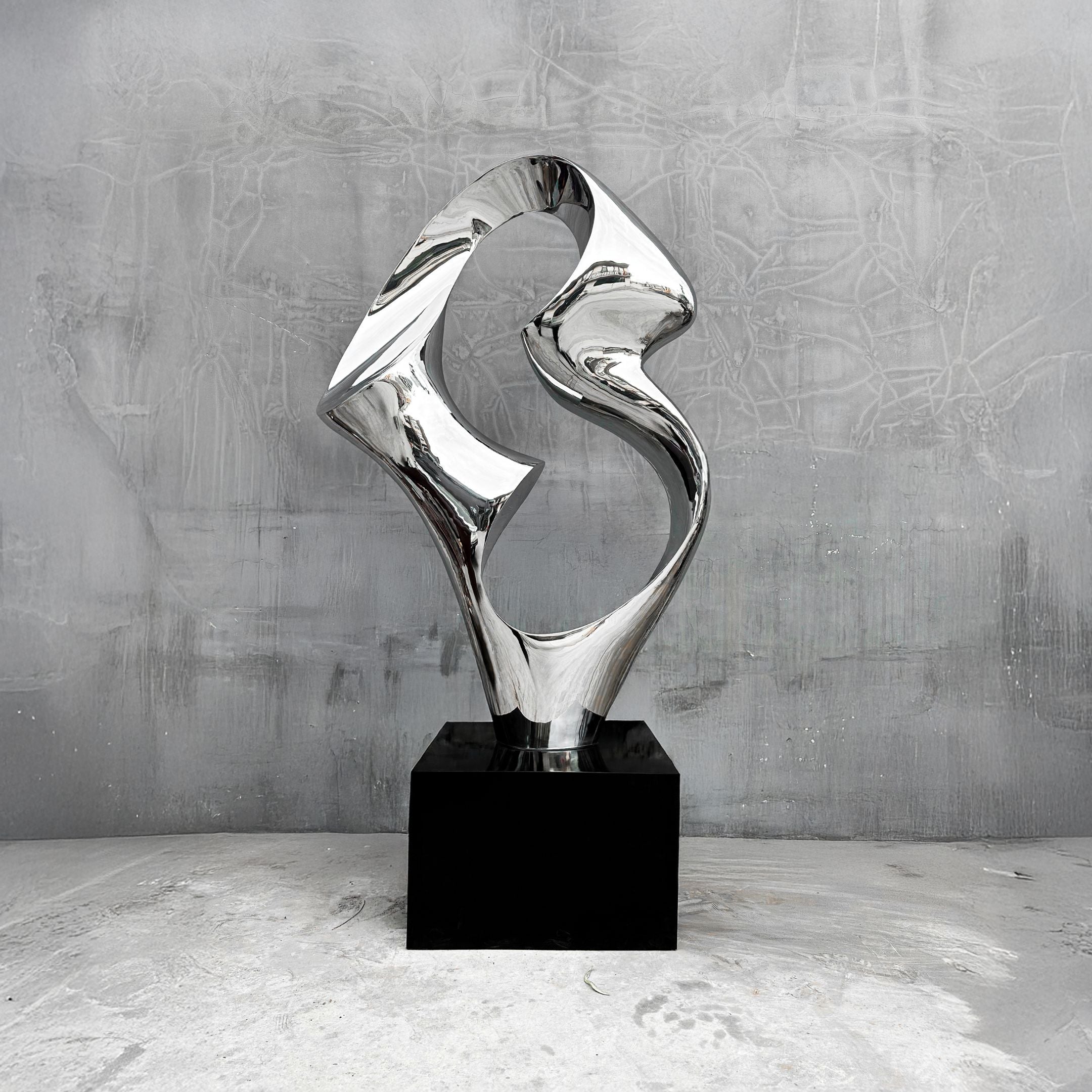 The Flow III Silver Organic Steel Sculpture by Giant Sculptures features smooth, twisting shapes on a black rectangular pedestal. Its abstract design against a textured gray wall and floor enhances its reflective surface and weather-resistant qualities.