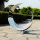 The Crescent Abstract Steel Outdoor Sculpture by Giant Sculptures is a shiny stainless steel piece featuring wave-like engravings. At 120 cm, it reflects surrounding greenery and sky as it stands on a patterned stone pathway, framed by lush bushes and tall trees.