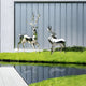 Two Noble Deer II Geometric Steel Sculptures, crafted from stainless steel and standing 198cm tall, are positioned on lush green grass beside a reflective water surface. In the background, a modern building featuring vertical glass panels adds to the scene's serene and contemporary vibe. These sculptures are part of the collection by Giant Sculptures.